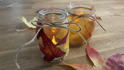 Teelichter in Gläsern mit bunten Herbstblättern dekoriert, auf einem Holzuntergrund platziert.