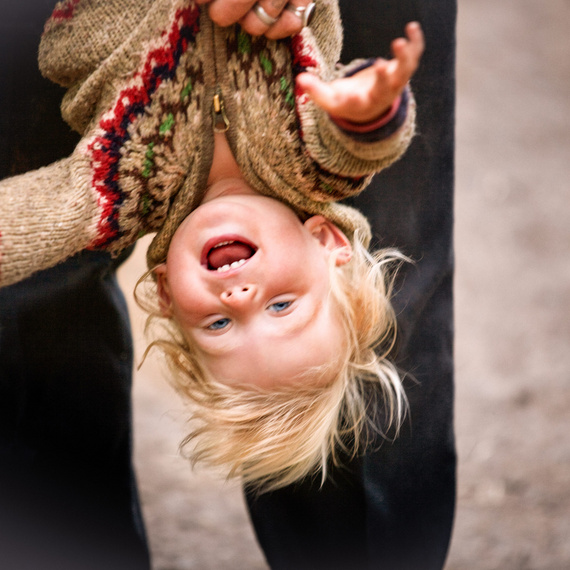 Lachendes Kind mit blonden Haaren, das auf dem Kopf hängend von einer erwachsenen Person gehalten wird, draußen auf unebenem Untergrund.