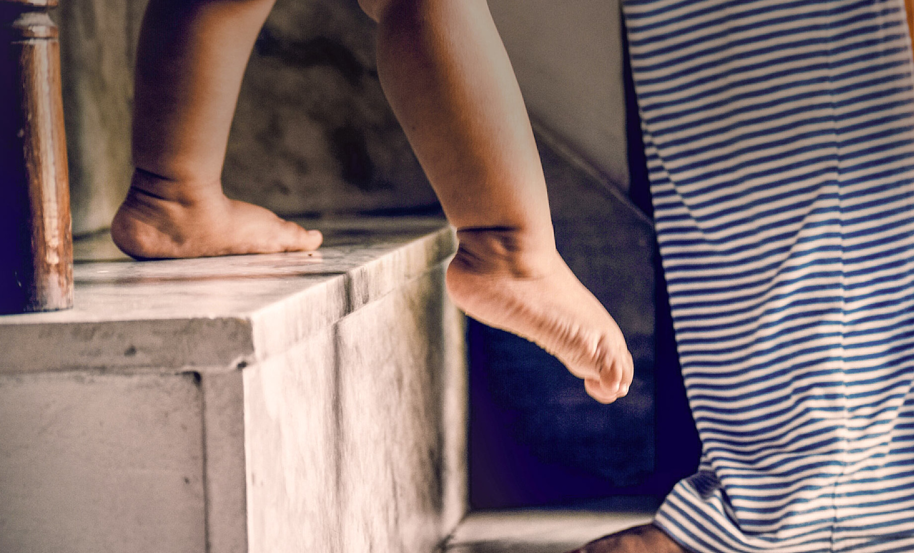 Kind, das auf eine Treppenstufe klettert, während ein Erwachsener mit gestreiften Hosen daneben steht.