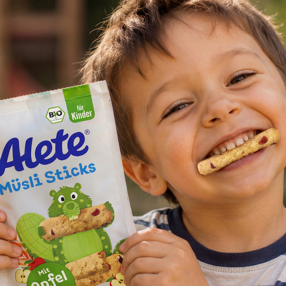 AI-generated Bild: Lächelnder Junge mit Alete Müsli Sticksl in der Hand und einem ausgepackten Stick im Mund, vor unscharfem Hintergrund eines Spielplatzes.