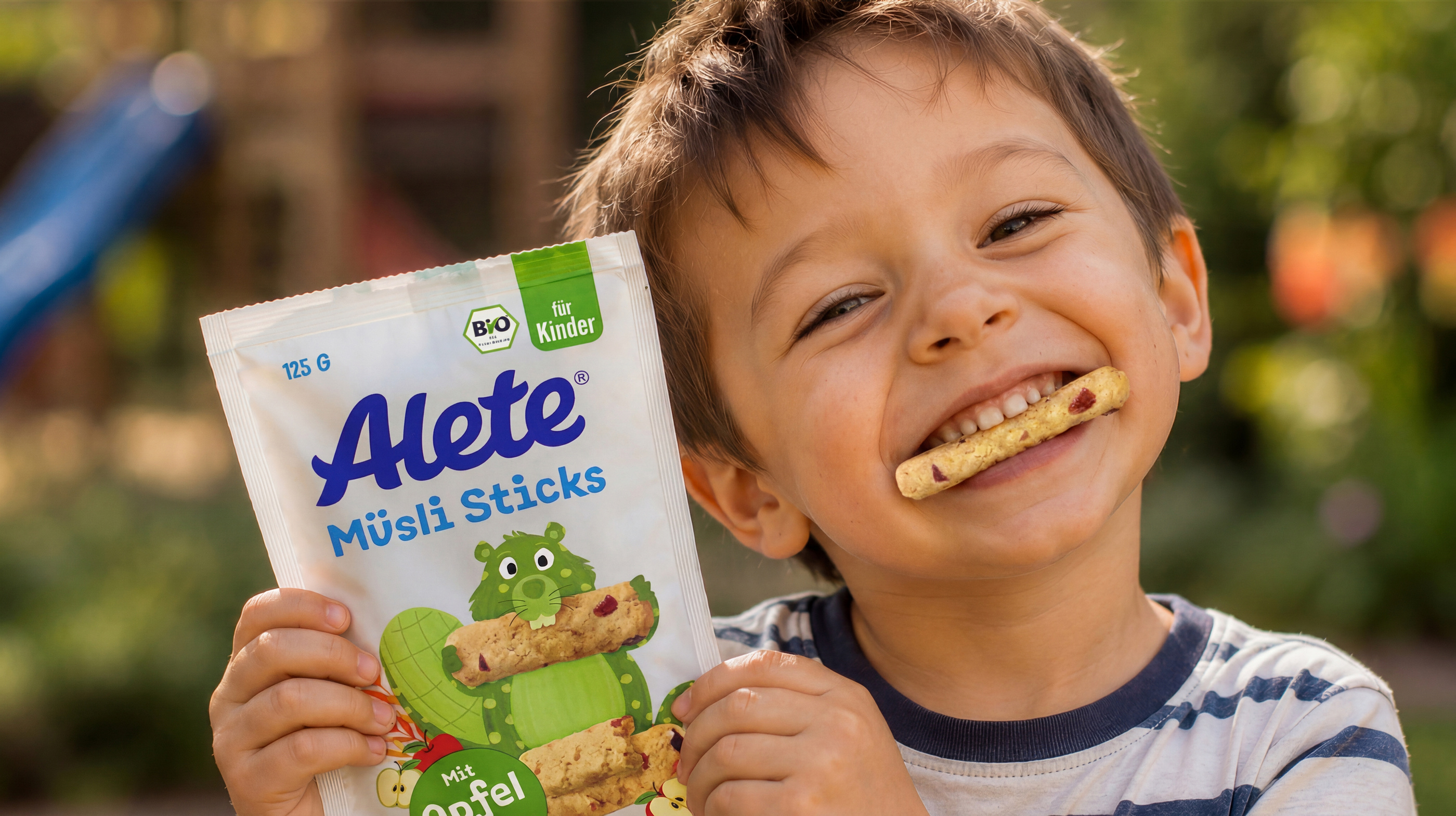 Lächelnder Junge mit Alete Müsli Sticksl in der Hand und einem ausgepackten Stick im Mund, vor unscharfem Hintergrund eines Spielplatzes.