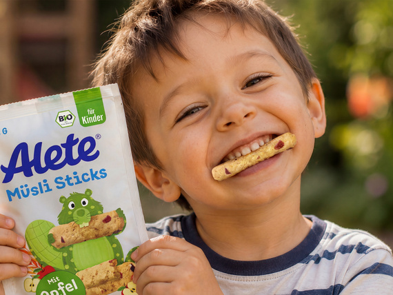 Lächelnder Junge mit Alete Müsli Sticksl in der Hand und einem ausgepackten Stick im Mund, vor unscharfem Hintergrund eines Spielplatzes.
