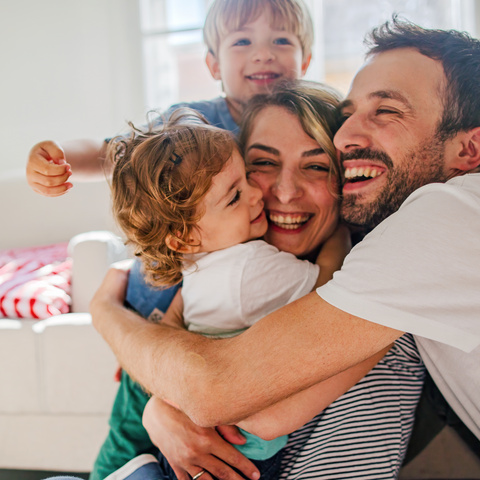 Familie umarmt sich lachend in einem heller eingerichteten Raum. Kinder zeigen Freude und Zuneigung.