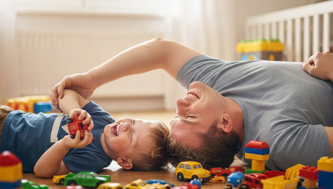 Vater und Sohn liegen lachend auf dem Boden, umgeben von bunten Spielzeugautos und Bauklötzen.
