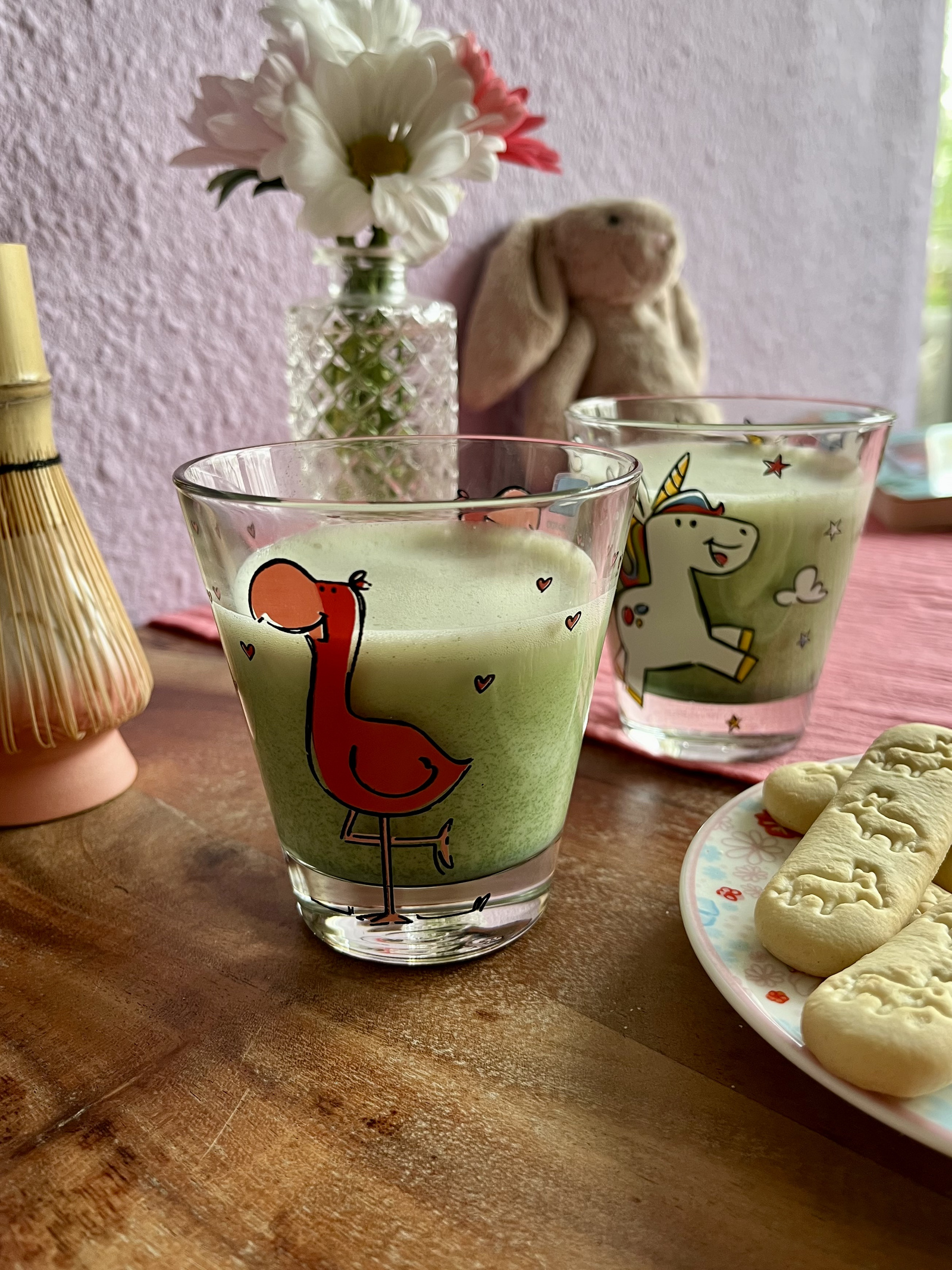 Bunte Gläser mit grünen Getränken, eines mit Flamingo-Design, dekoriert mit Blumen und Keksen auf einem Tisch.
