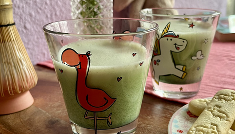 Bunte Gläser mit grünen Getränken, eines mit Flamingo-Design, dekoriert mit Blumen und Keksen auf einem Tisch.