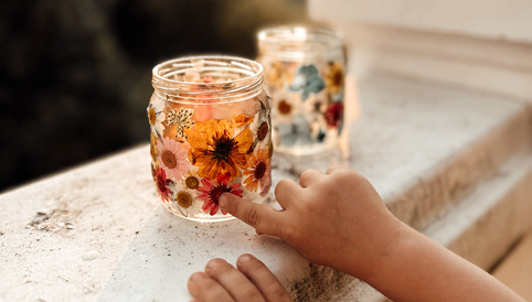 Ein Kind berührt sanft ein dekoratives Glas mit getrockneten Blumen, das auf einem Balkongeländer steht, während die Sonne untergeht.