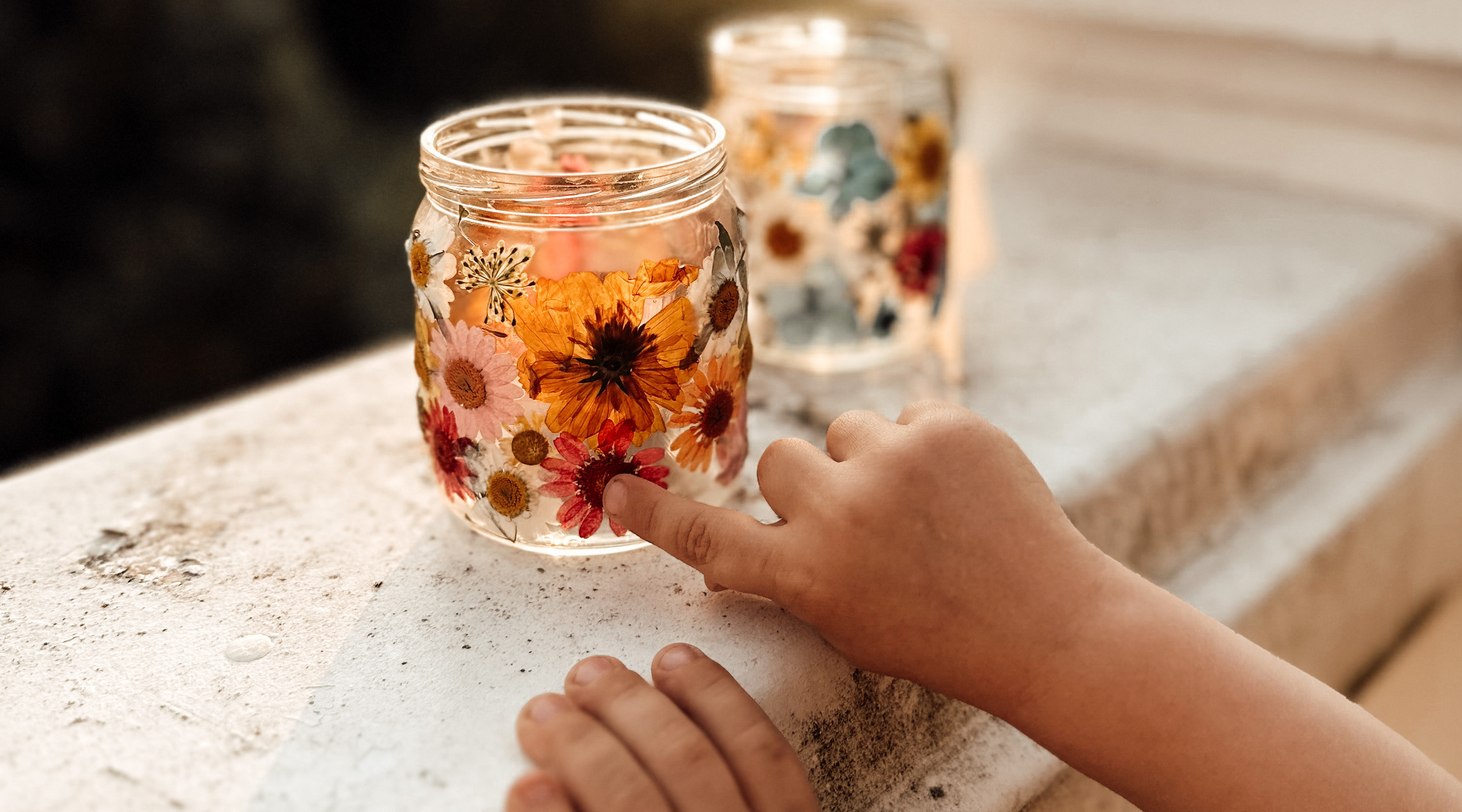 Ein Kind berührt sanft ein dekoratives Glas mit getrockneten Blumen, das auf einem Balkongeländer steht, während die Sonne untergeht.
