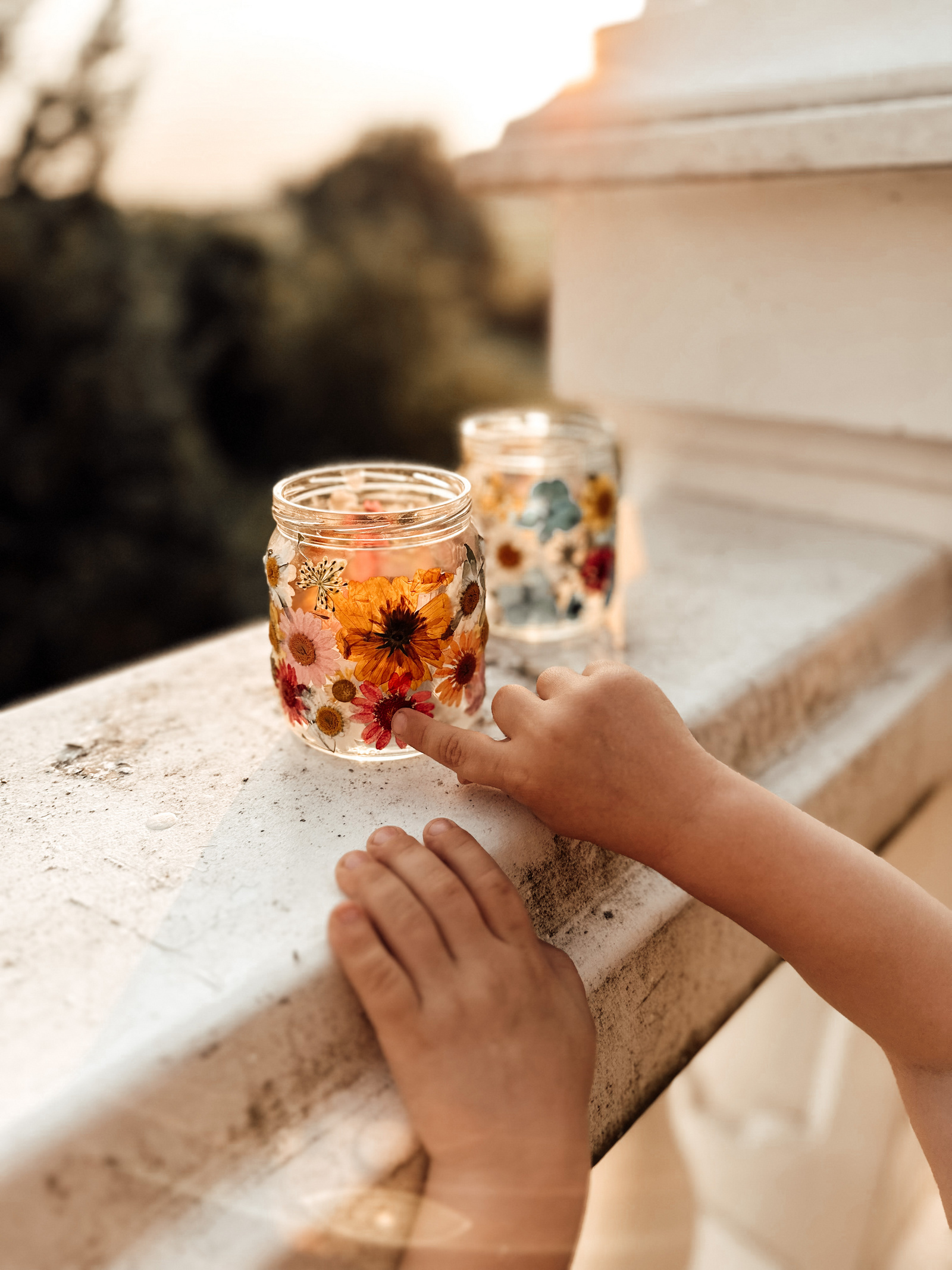 Ein Kind berührt sanft ein dekoratives Glas mit getrockneten Blumen, das auf einem Balkongeländer steht, während die Sonne untergeht.