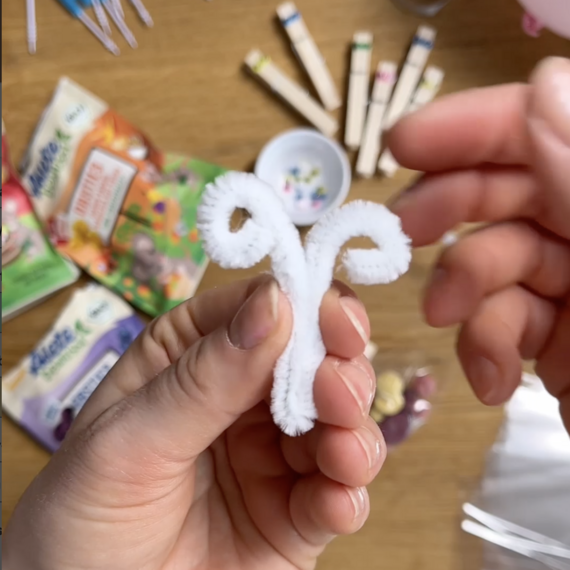 Bastelprojekt: Hand hält eine weiße Pfeifenreinigerfigur in Form eines Herzes, umgeben von Bastelmaterialien.