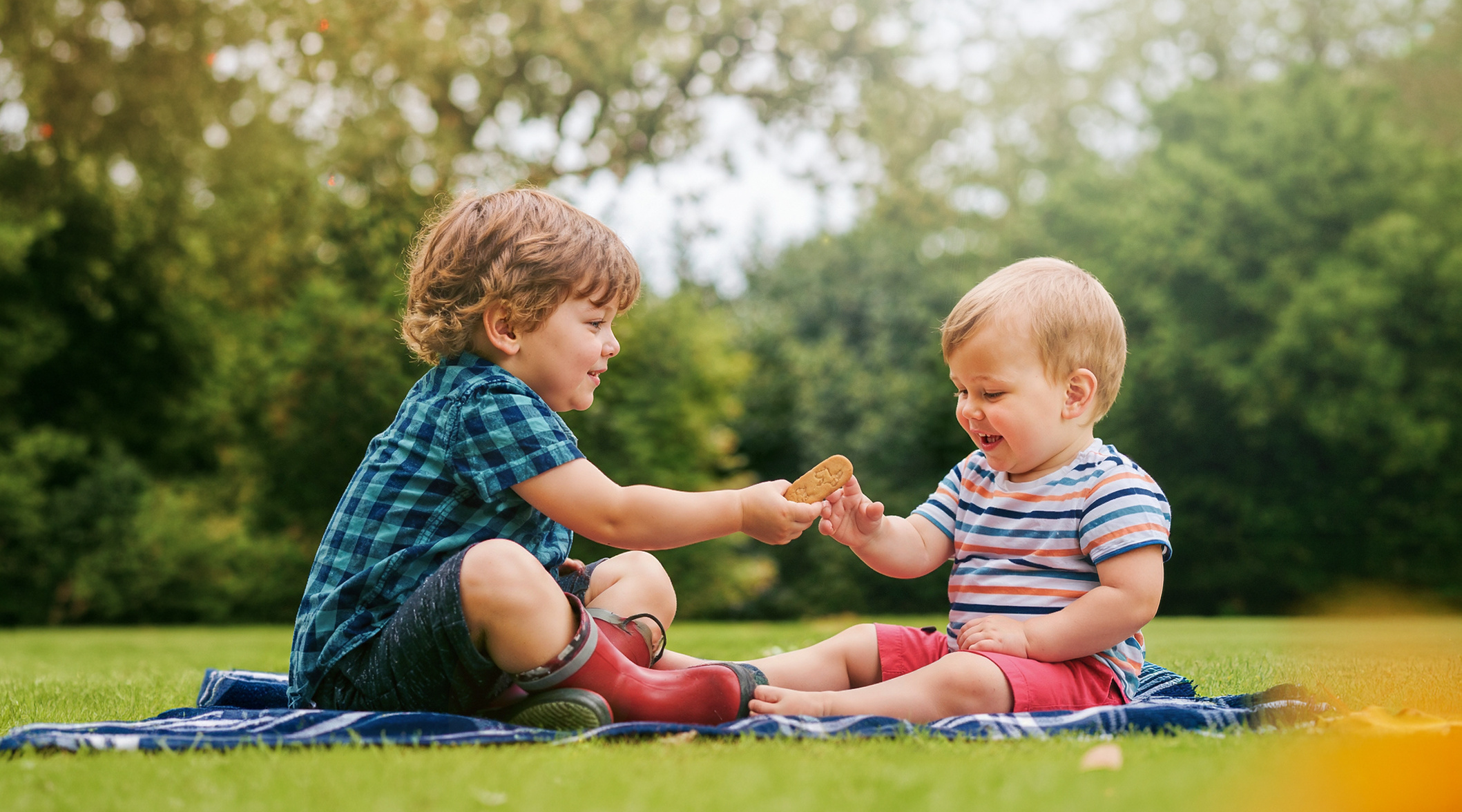 Zwei kleine Kinder im Freien, die auf einer Decke sitzen und zusammen Alete Kinderkekse teilen. Der große Junge reicht dem jüngeren Bruder einen Keks, während dieser lächelt.
