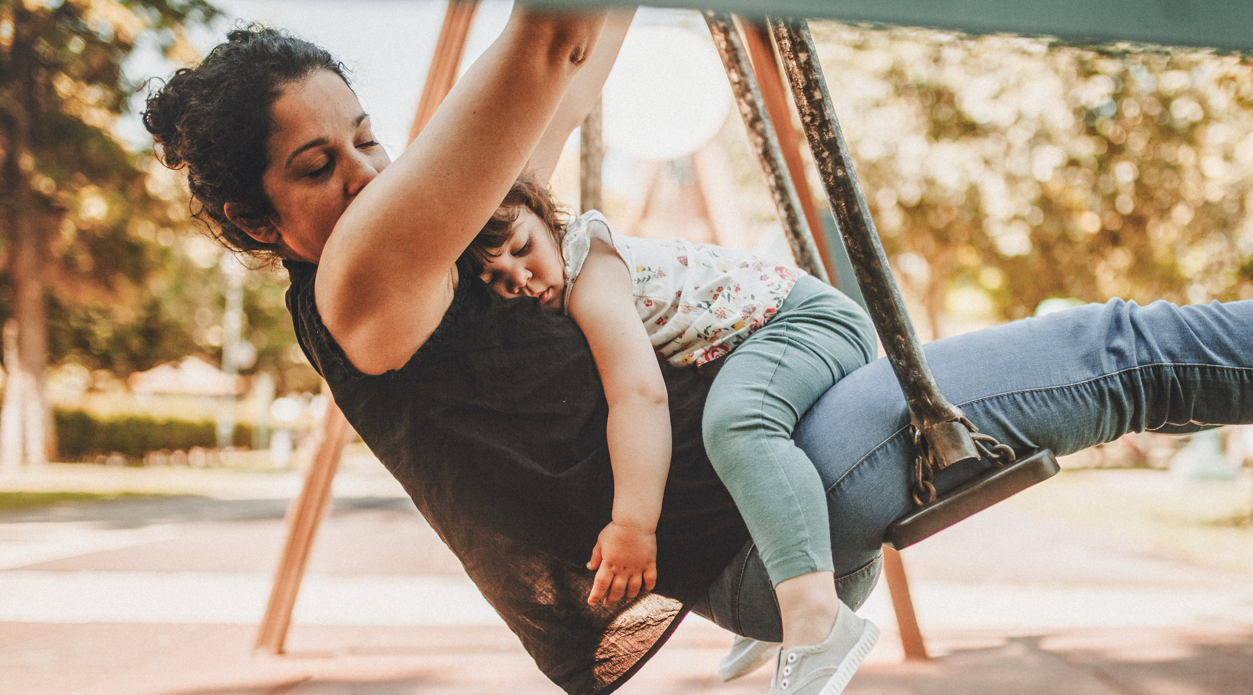 Mutter schwingt mit schlafendem Kind auf einer Schaukel im Park, umgeben von Bäumen und natürlichem Licht.