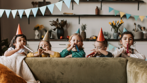 Fünf fröhliche Kinder mit Partyhüten blasen auf Musikinstrumenten und feiern in einem festlich dekorierten Raum.