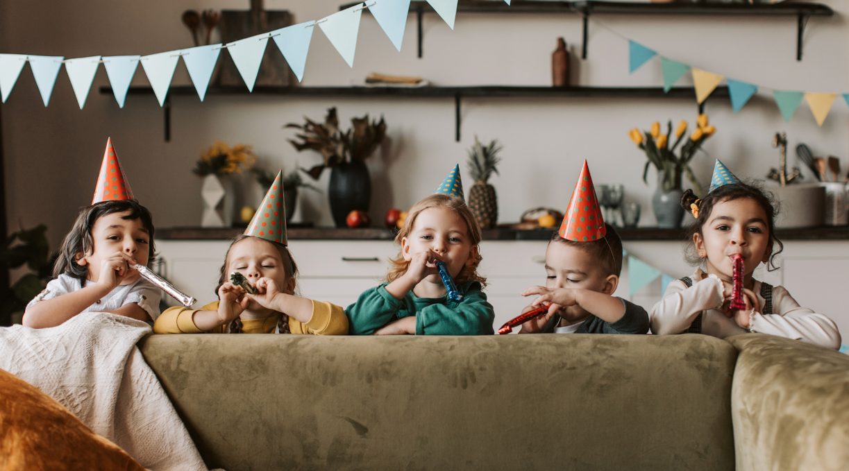 Fünf fröhliche Kinder mit Partyhüten blasen auf Musikinstrumenten und feiern in einem festlich dekorierten Raum.