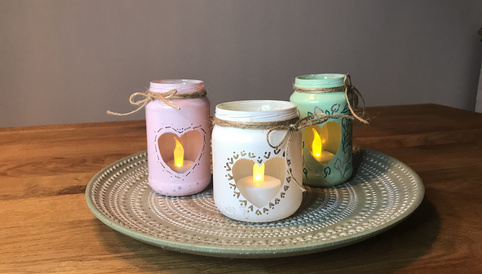 Dekorative Gläser mit Herzen, in pink, weiß und grün, auf einem stilvollen Teller, angezündet mit LED-Kerzen für gemütliche Atmosphäre.