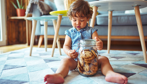 Kleines Kind mit lockigen Haaren sitzt auf einem Teppich und greift in ein Glas mit Keksen. Im Hintergrund ist ein modernes Wohnzimmer sichtbar.