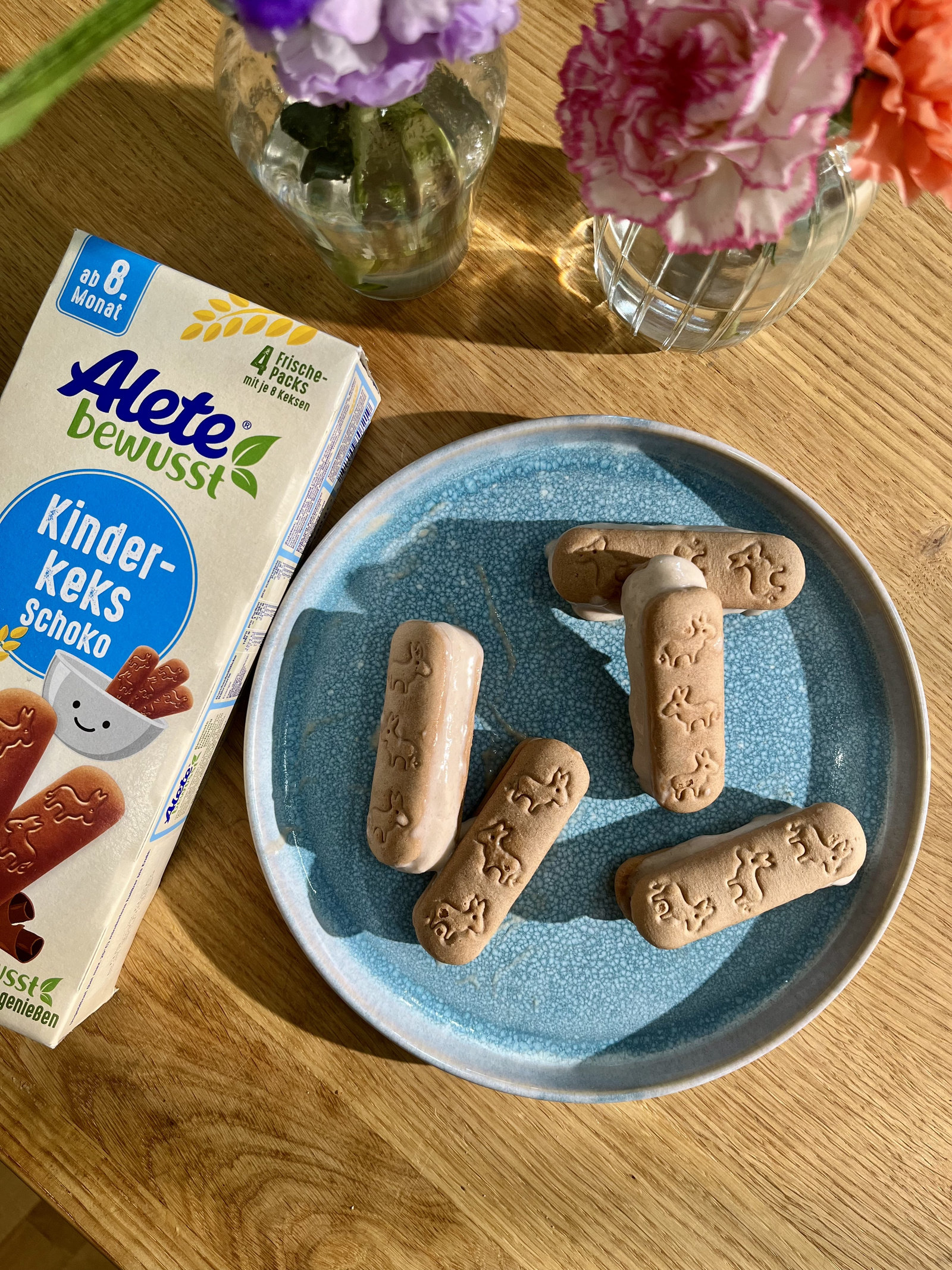 Alete Kinderkekse mit Schokolade auf einem blauen Teller, umgeben von bunten Blumen in Glasvasen. Ideal für Babys ab 8 Monaten.