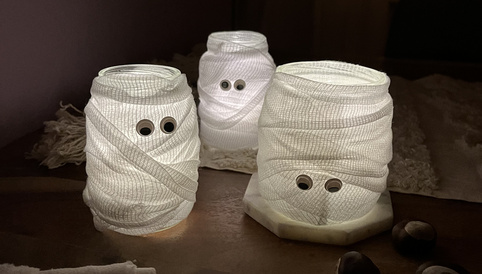 Leuchtende Geister aus Alete-Gläschen, umwickelt mit Bandagen und mit googly Augen verziert, auf einem Holztablett.