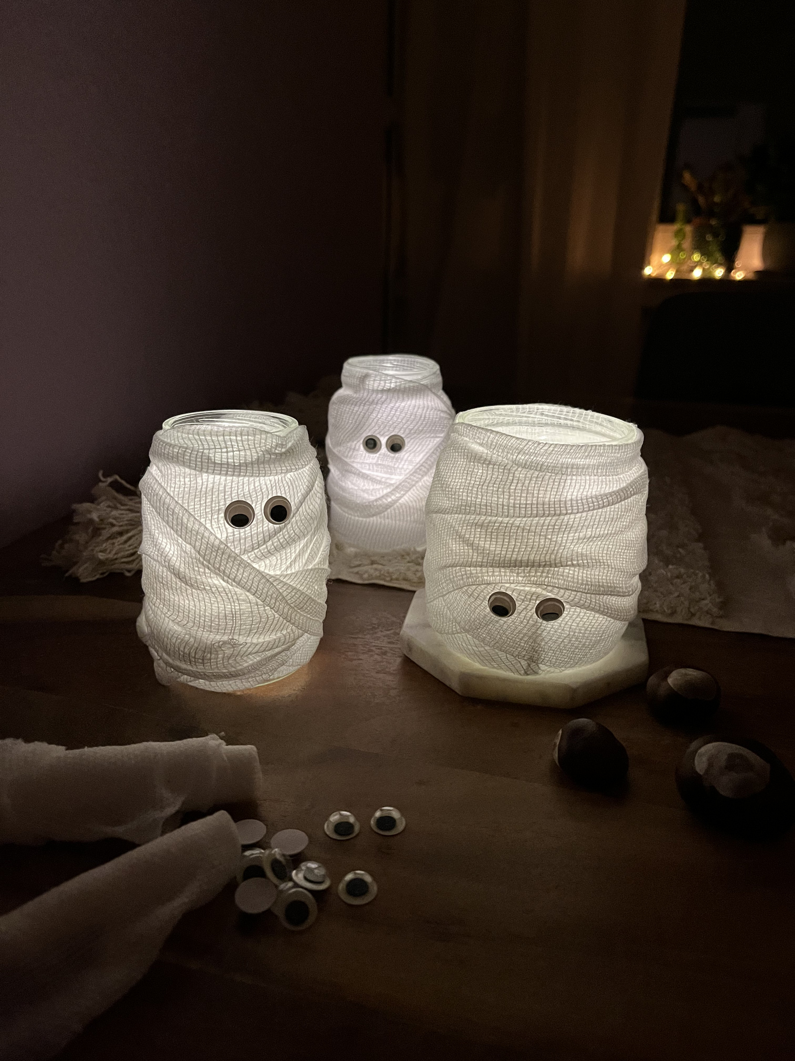 Leuchtende Geister aus Alete-Gläschen, umwickelt mit Bandagen und mit googly Augen verziert, auf einem Holztablett.