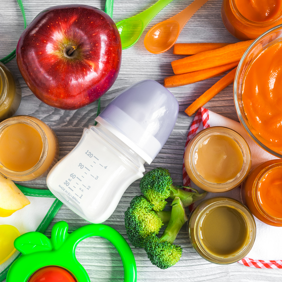 Bunte Auswahl an Baby-Nahrungsmitteln: Apfel, Möhren, Brokkoli, Gläser mit Brei und eine Babyflasche auf einem Holztisch.