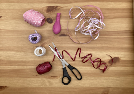 Bastelmaterialien auf einem Holztisch: Bänder in Pink- und Lilatönen, eine pinke Ballonform, Schere und Klebeband.