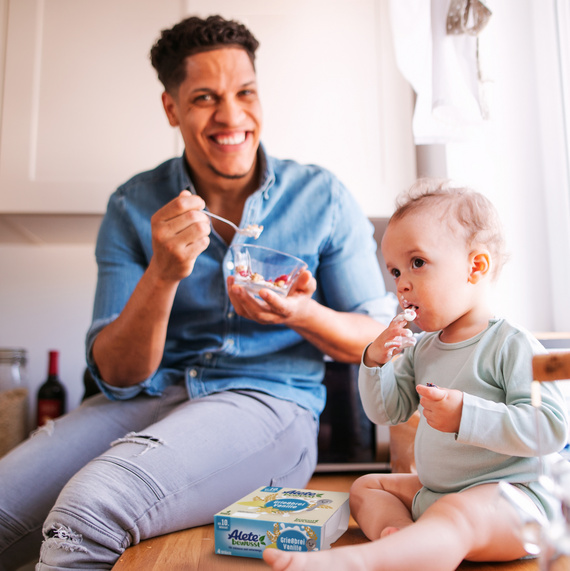 Vater und Kleinkind genießen ein gemeinsames Frühstück in der Küche, während der Vater aus einer Schüssel isst und das Kind ein Joghurt mit Löffel probiert.