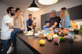 Familienmitglieder und Freunde stehen um eine moderne Kücheninsel, während sie gemeinsam ein Gericht zubereiten. Auf dem Tisch liegen frische Zutaten wie Gemüse und Milchprodukte.