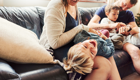 Familie entspannt auf einem Sofa; Mutter lacht und hält einen fröhlichen, blondhaarigen Jungen auf ihrem Schoß, während ein weiterer Junge neben ihnen sitzt.