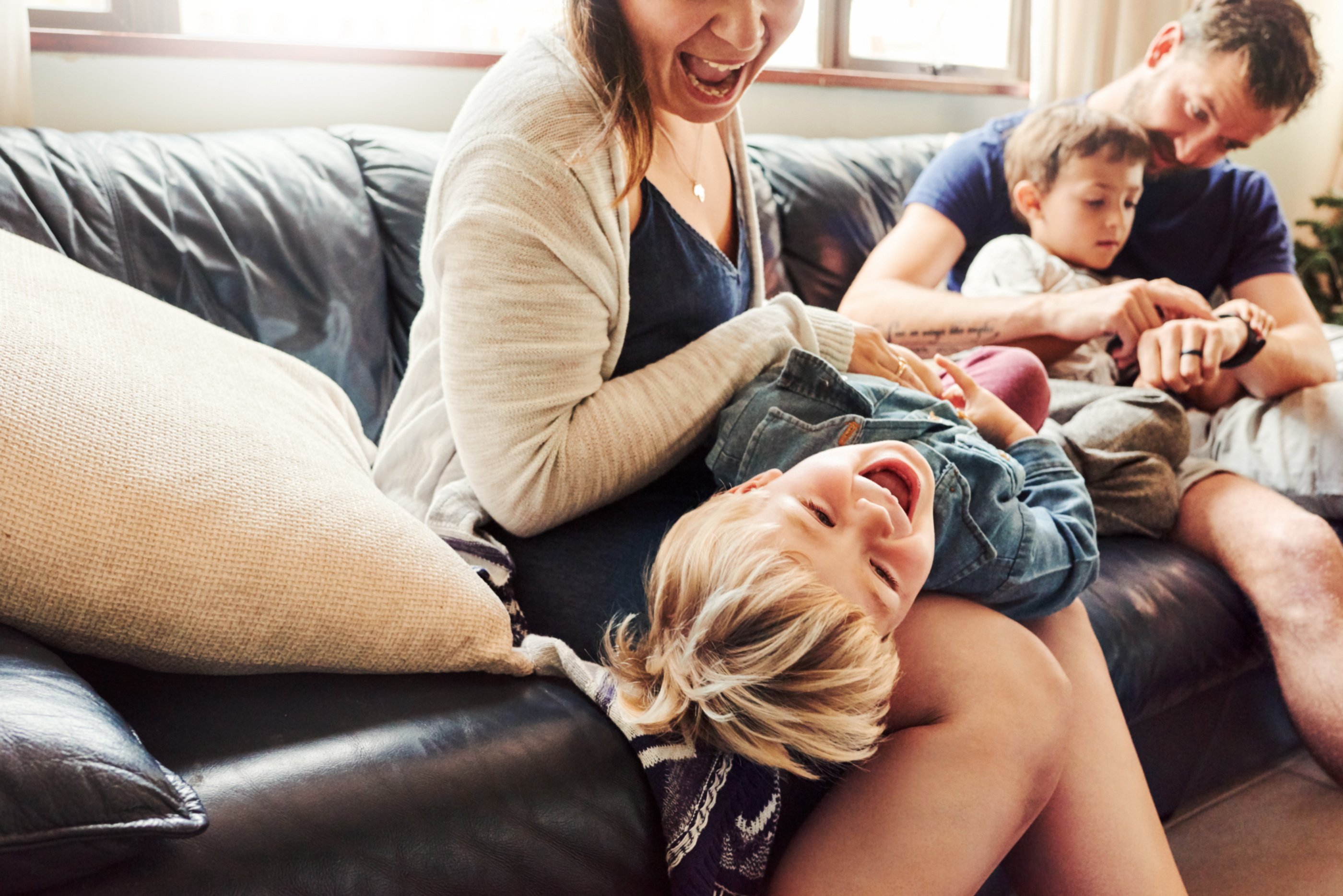 Familie entspannt auf einem Sofa; Mutter lacht und hält einen fröhlichen, blondhaarigen Jungen auf ihrem Schoß, während ein weiterer Junge neben ihnen sitzt.
