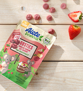 Alete Obsties: Bio Obstsnacks für Kinder mit Erdbeere und Banane, umgeben von Erdbeeren und runden Snacks auf Holzuntergrund.