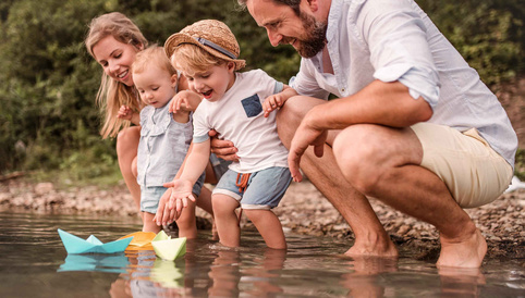 Familie am Ufer eines Gewässers, während Kinder mit bunten Papierbooten spielen und lachen.