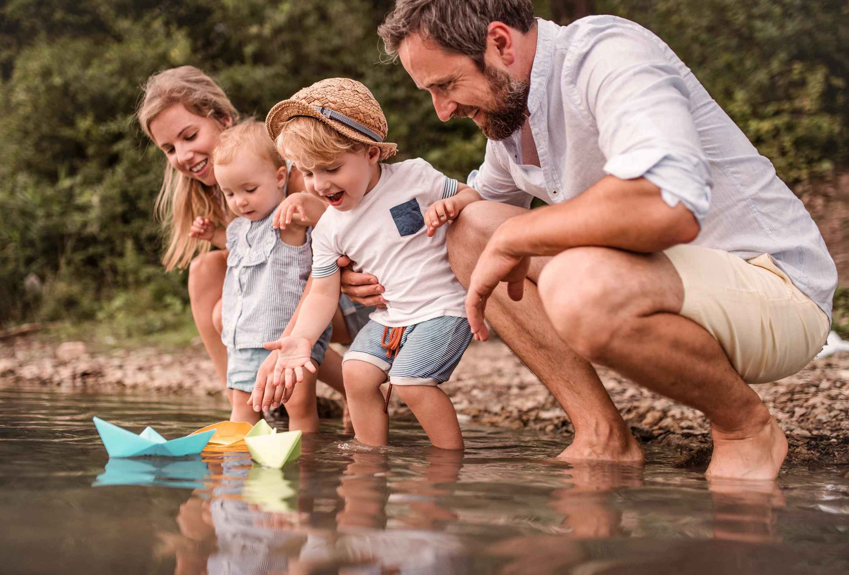 Familie am Ufer eines Gewässers, während Kinder mit bunten Papierbooten spielen und lachen.