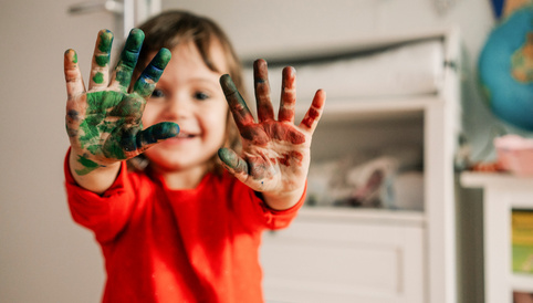Lächelndes Kind mit bemalten Händen in einem kreativen Spielumfeld. Farbflecken in Grün, Blau und Rot sichtbar.