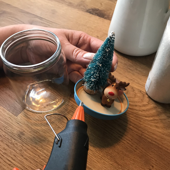 Kreative Bastelarbeit mit einer Heißklebepistole, Miniatur-Weihnachtsbaum und Rentierfigur auf einem Holztisch.