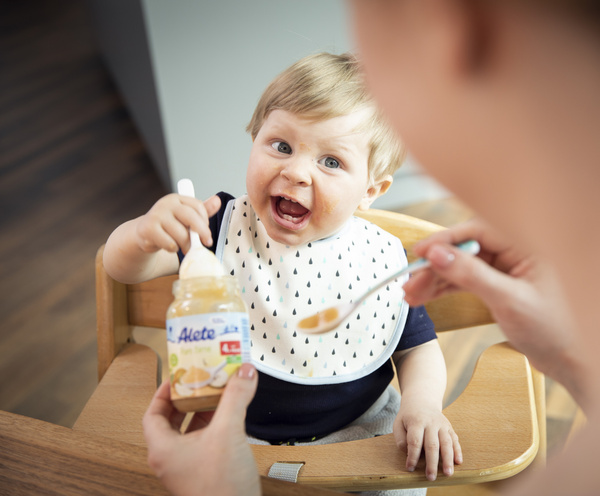 Lächelnder Säugling im Hochstuhl, der begeistert ein Glas Babynahrung hält und neugierig nach einem Löffel greift.