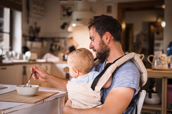 Vater füttert sein Baby, das in einem Tragetuch sitzt, am Küchentisch. Das Bild zeigt einen warmen, familiären Moment.