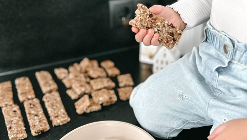 Selbstgemachte Müsliriegel auf einem Tisch, ein Kind hält einen Müsli-Riegel in der Hand, mehrere Riegel liegen daneben.
