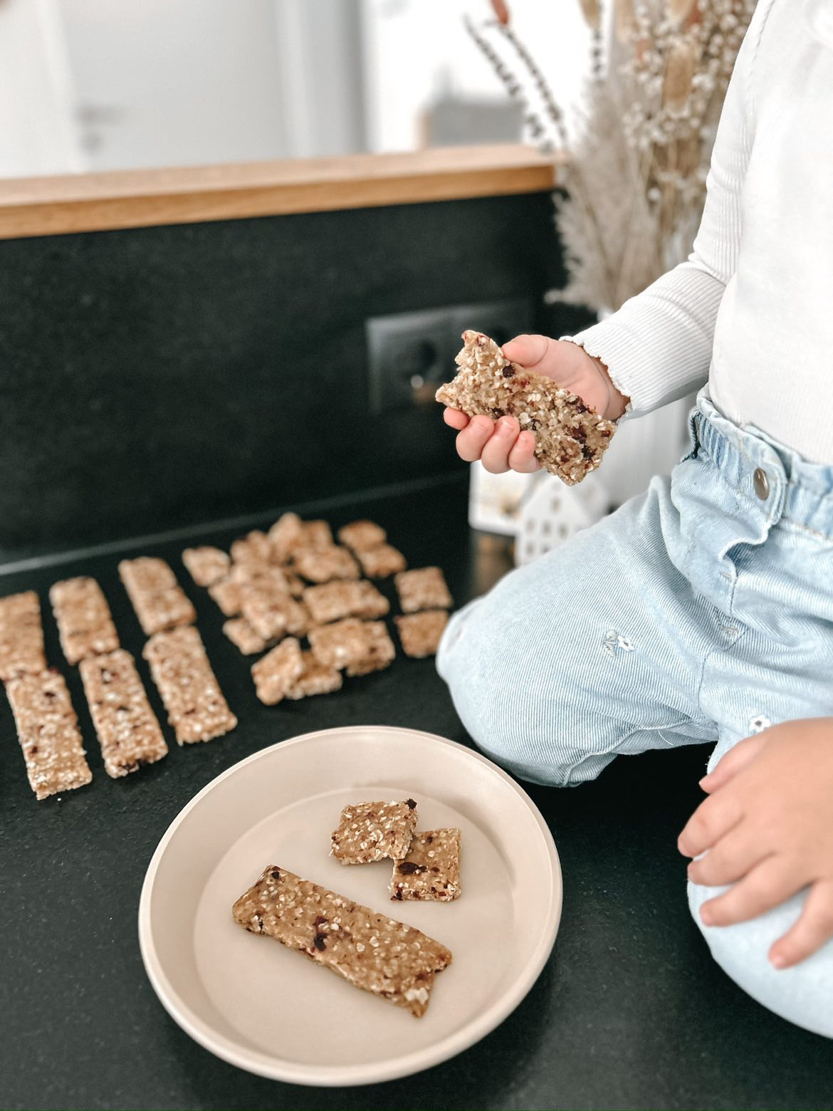 Selbstgemachte Müsliriegel auf einem Tisch, ein Kind hält einen Müsli-Riegel in der Hand, mehrere Riegel liegen daneben.