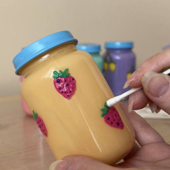 Hand, die mit einem Wattestäbchen eine dekorierte Glasflasche mit Erdbeermotiven bemalt, im Hintergrund farbige Flaschen.