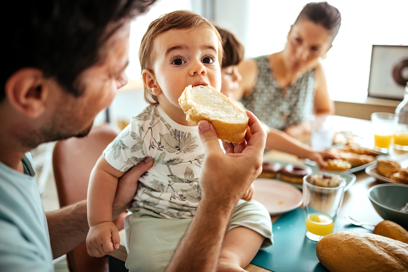 Kleines Kind beim Essen von Brot mit Frischkäse, während es von einem Erwachsenen auf dem Schoß gehalten wird. Frühstückstisch mit verschiedenen Speisen im Hintergrund.