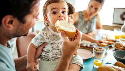 Kleines Kind beim Essen von Brot mit Frischkäse, während es von einem Erwachsenen auf dem Schoß gehalten wird. Frühstückstisch mit verschiedenen Speisen im Hintergrund.