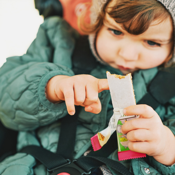 Kleines Kind in warmer Jacke hält einen Alete-Riegel und betrachtet ihn aufmerksam.