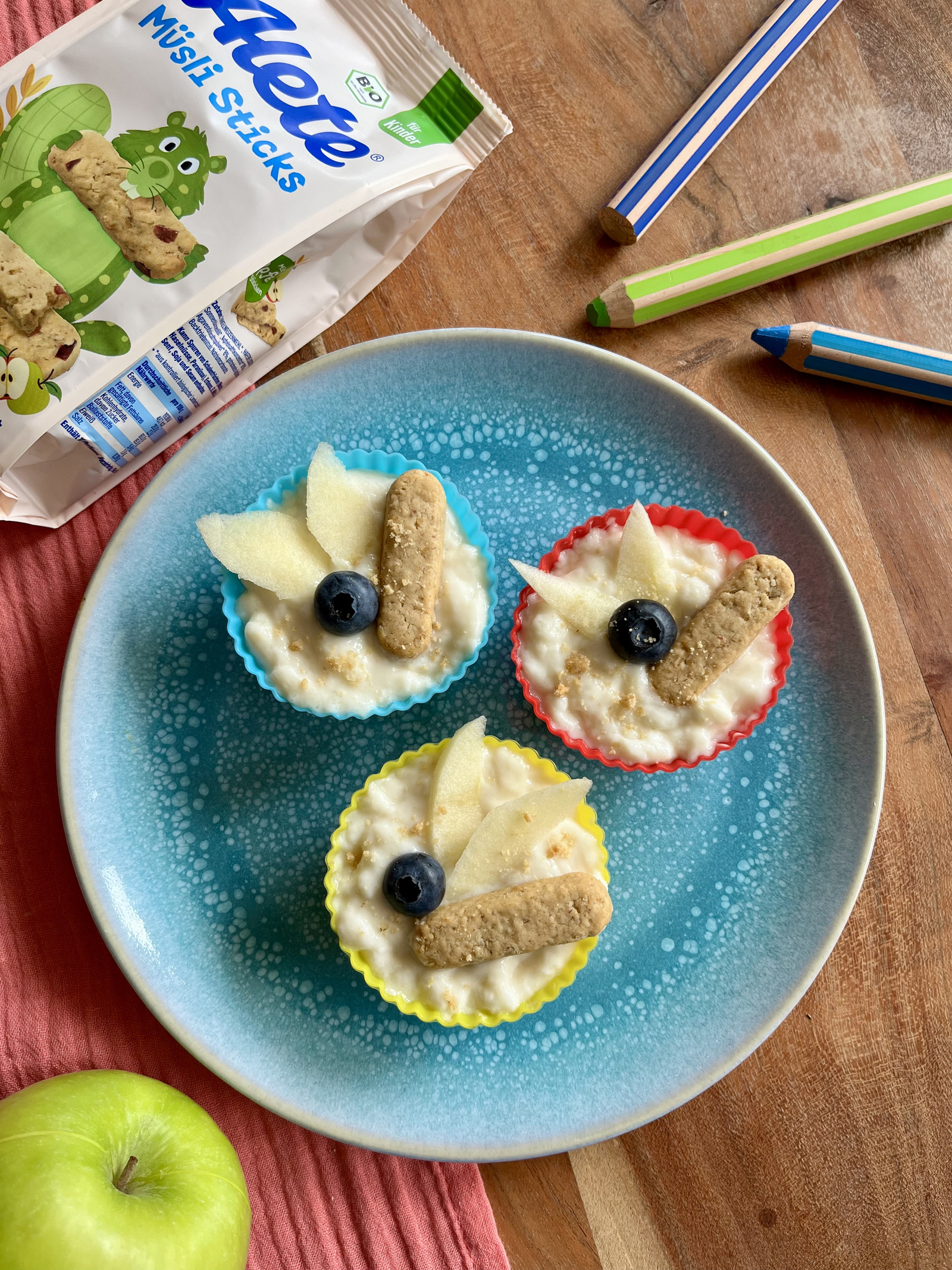 Muffins in bunten Förmchen mit Birnenscheiben, Heidelbeeren und Alete Müslisticks auf blauem Teller.
