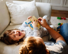 AI-generated Bild: Lachendes Kind auf dem Sofa hält eine Packung Alete Dinkelkeks, während ein Erwachsener ihm hilft. Spielzeug liegt im Hintergrund.