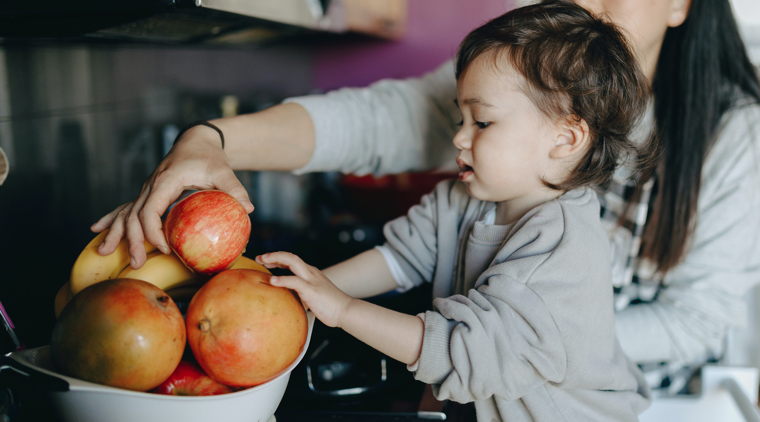Kleinkind greift nach roten Äpfeln aus einer Schüssel mit frischem Obst, während eine Hand im Hintergrund assistiert.