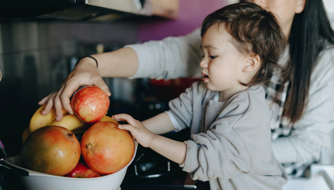 Kleinkind greift nach roten Äpfeln aus einer Schüssel mit frischem Obst, während eine Hand im Hintergrund assistiert.