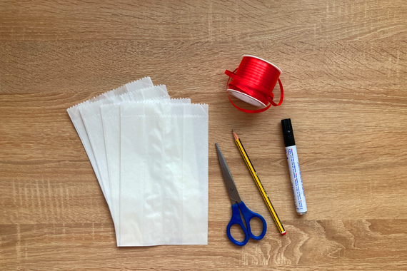 Materialien zum Basteln: weiße Papierbeutel, Schere, rotes Band, Bleistift und Marker auf Holzuntergrund.