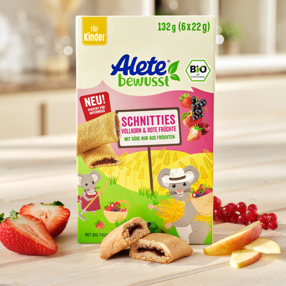 Verpackung von Alete Schnitties mit Vollkorn und roten Früchten, abgebildet mit frischen Erdbeeren und Apfelscheiben. Ideal für Kinder und Bio-qualifiziert.