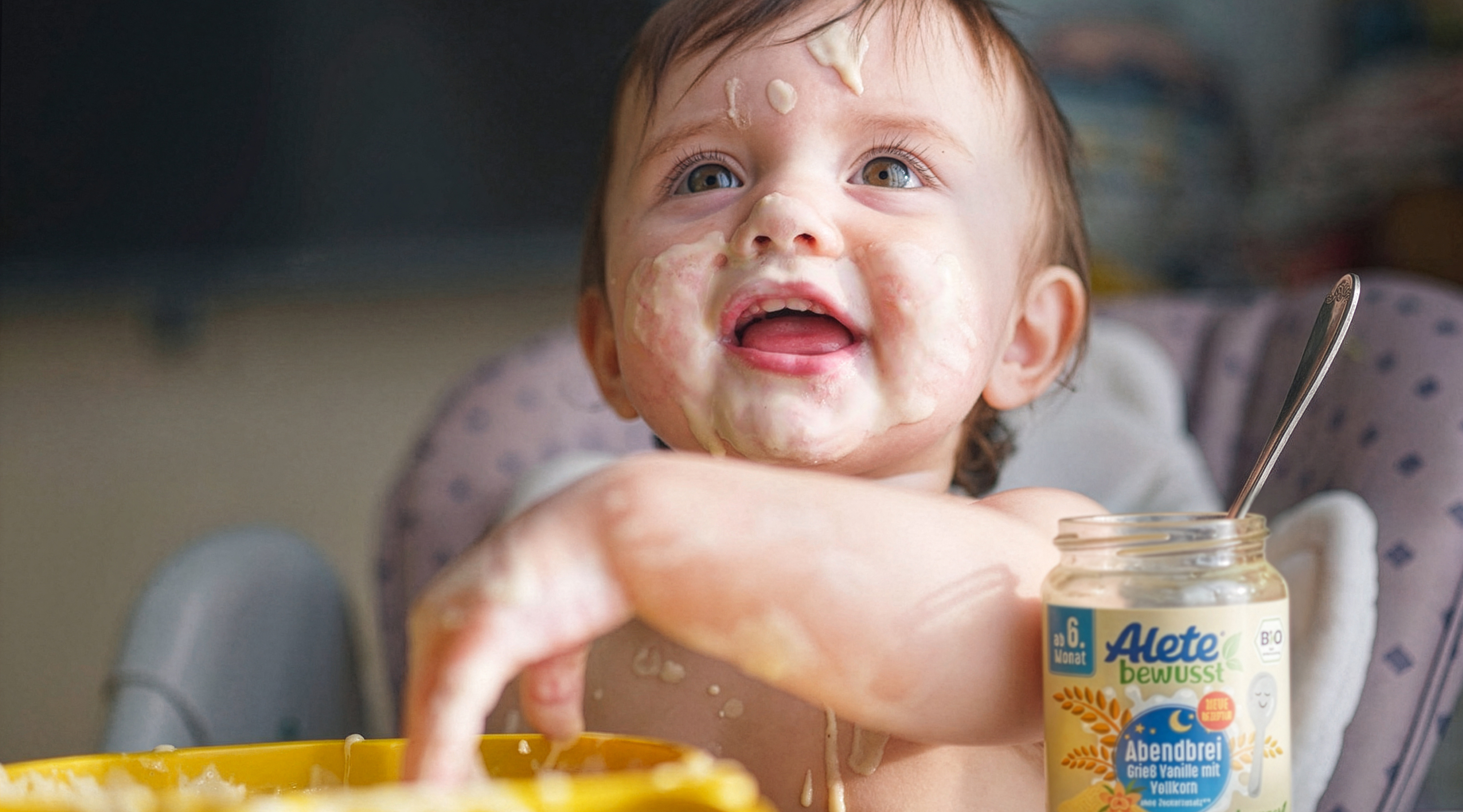 Lächelndes Baby mit Essensreste im Gesicht, das aus einem gelben Schälchen isst, neben einem Glas Alete Abendbrei.