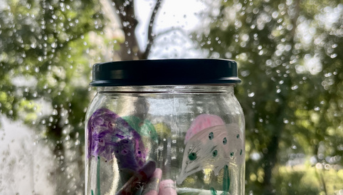 Bunte, handgeschriebene Zettel in einem Glas, gehalten vor einem regennassen Fenster mit unscharfem Hintergrund.